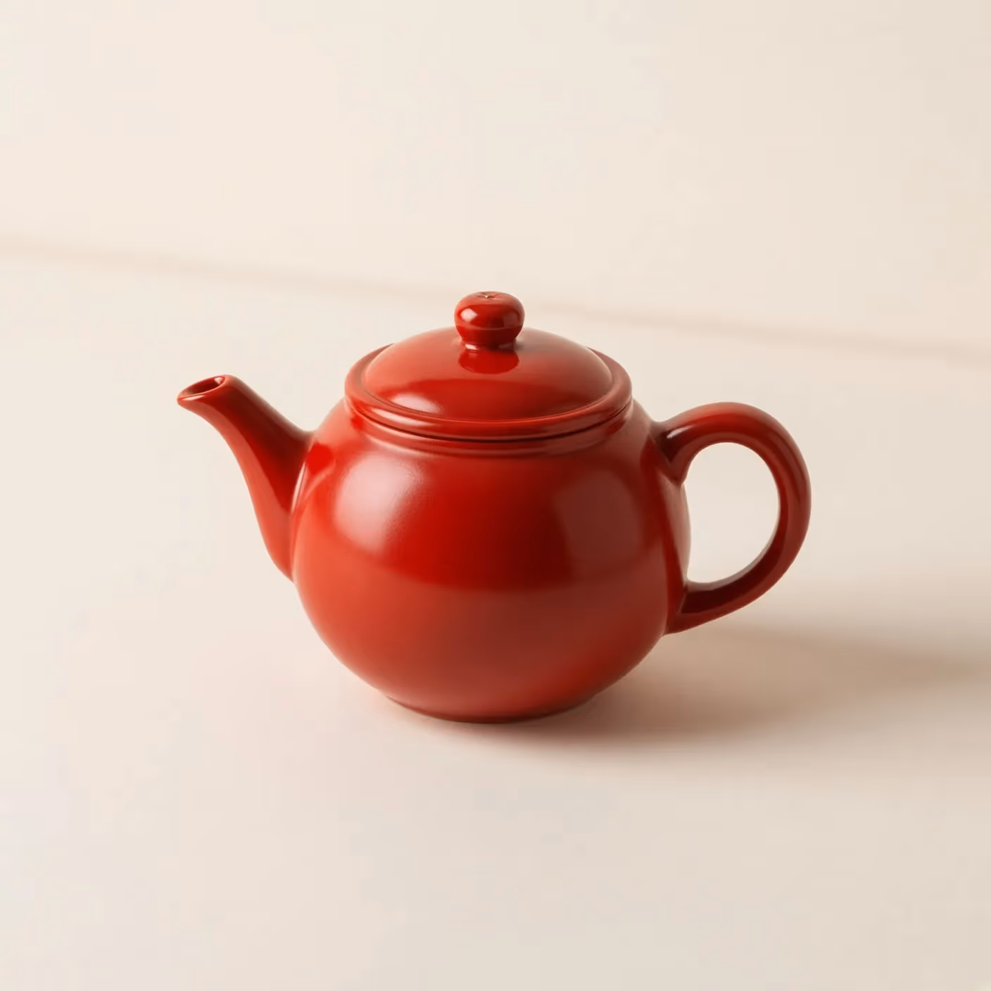 A small red ceramic teapot on a plain cream table, soft daylight, clean studio photograph, no lettering.